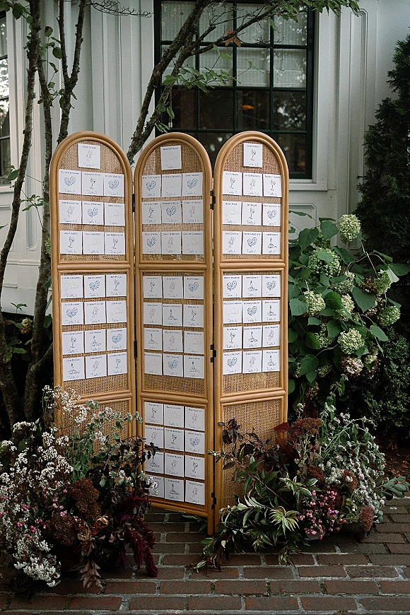 Wedding seating chart seating chart display on a rattan folding screen with hydrangeas and greenery along a brick walkway by a house exterior