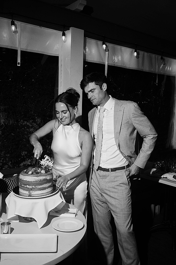Wedding cake cutting as the bride cuts wedding cake with a knife and server while the groom watches under string lights by windows at night