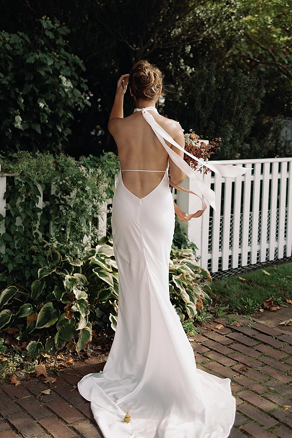 Bridal portrait of a bride in a backless wedding dress holding a bouquet, seen from behind on a brick path by a white fence and greenery