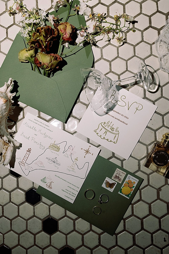 Wedding invitation suite with custom illustrated wedding invitation, green envelopes, rings, perfume, and dried flowers on a hexagon tile floor