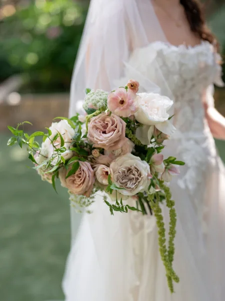 Bridal bouquet of blush and ivory roses, ranunculus, and greenery with trailing amaranthus, held against a lace dress and veil in garden bokeh
