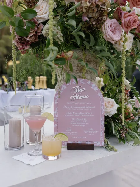 Wedding bar menu sign with calligraphy listing signature cocktails beside rose and hydrangea florals on a white linen bar table outdoors