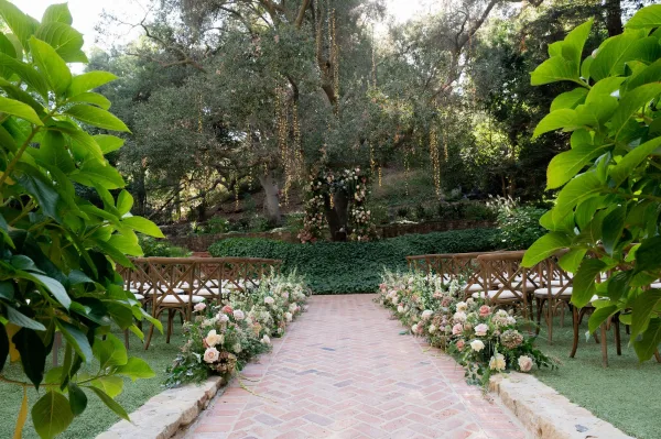 Ceremony aisle design with blush and white florals lining a brick walkway, leading to a floral arch under string lights in a garden setting