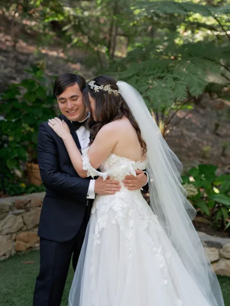 Couple portrait of bride and groom embrace, her long veil and floral hairpiece against garden greenery with a stone wall backdrop