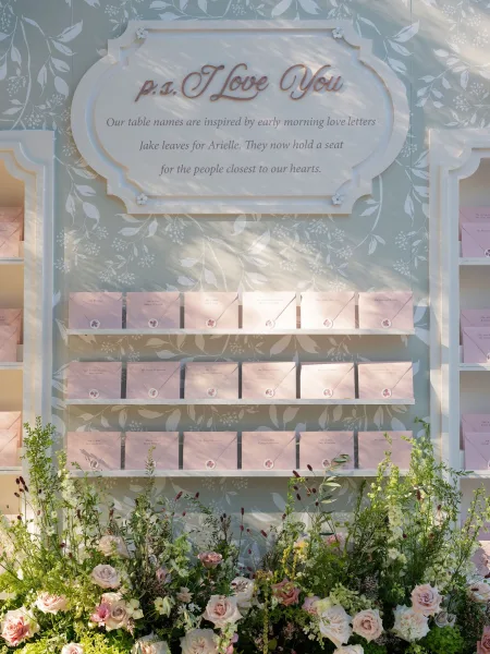 Escort card display with wedding escort cards in soft pink envelopes, wax seals, and roses on shelving against a floral backdrop in sun shadows