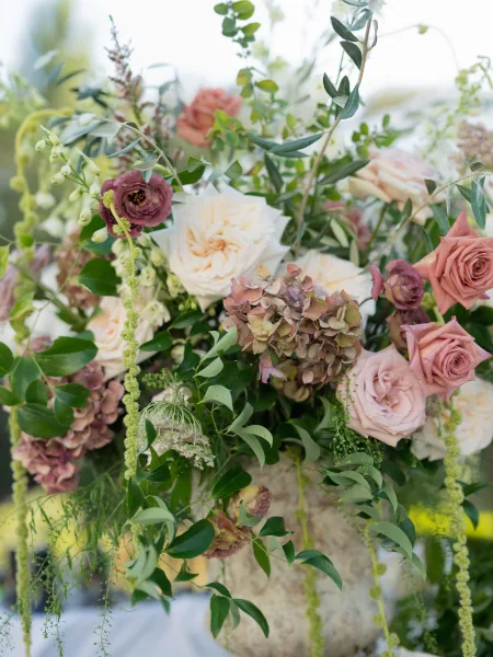 Wedding floral arrangement in a floral urn with roses, hydrangea, and hanging amaranthus, set on a white-linen table in a blurred garden backdrop