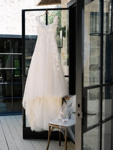Wedding dress with lace bodice hanging on a black-framed glass door, tulle skirt falling beside a chair with white shoes