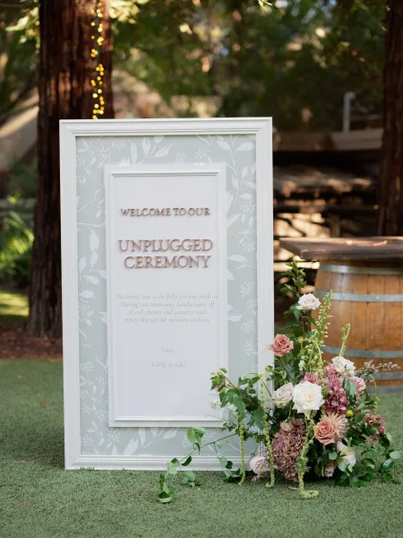 Wedding welcome sign in a white frame with roses and hydrangea greenery on a wooden barrel beneath string lights in a garden lawn setting