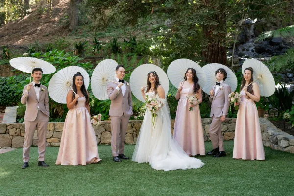 Wedding party portrait with paper parasols, bride in veil and bouquet flanked by groomsmen in tan suits in a garden by a waterfall