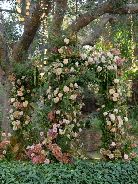 Wedding ceremony arch with roses and hydrangeas, greenery and string lights, set under oak trees by a stone garden wall
