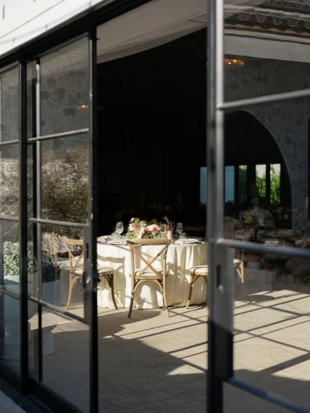 Reception tablescape with ivory linen, pastel floral centerpiece, wine glasses and place settings, sunlit by glass doors against stone walls