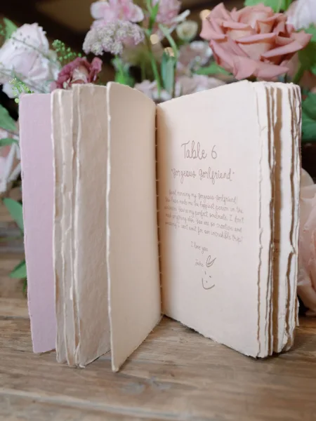 Wedding table numbers in a table number book with handwritten calligraphy on deckled edge paper, accented by pink roses on a wood table
