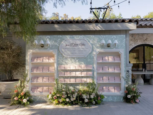 Wedding seating chart with pastel escort card display on built-in shelves, floral and greenery accents, beneath string lights on a stone patio wall