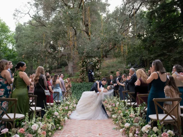 Ceremony kiss at an outdoor wedding ceremony, groom dipping bride in flowing cathedral veil on a brick aisle under string lights and floral arch