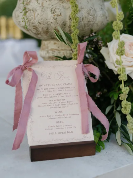 Bar menu sign with wedding bar menu details, pink ribbon bows and calligraphy on a stand beside a white rose on a linen table outdoors