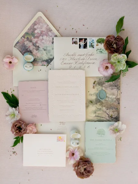 Invitation suite wedding invitation flat lay with embossed cards, calligraphy envelopes, wax seal, postage stamps, rings, and flowers on linen backdrop