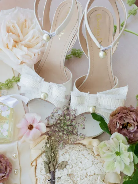 Bridal shoes with white bow wedding heels and pearl details beside flowers, lace ring pillow, and perfume bottle on neutral fabric