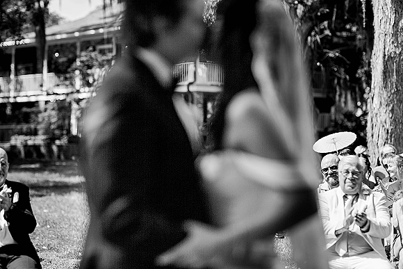 Ceremony moment at an outdoor wedding ceremony as guests applaud, bride in gown and groom in tux on a lawn by a house veranda with a sun parasol