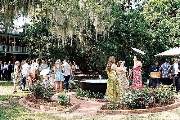 Wedding cocktail hour guests mingling at an outdoor wedding cocktail hour bar on brick pavers under oak trees with hanging moss and a white parasol
