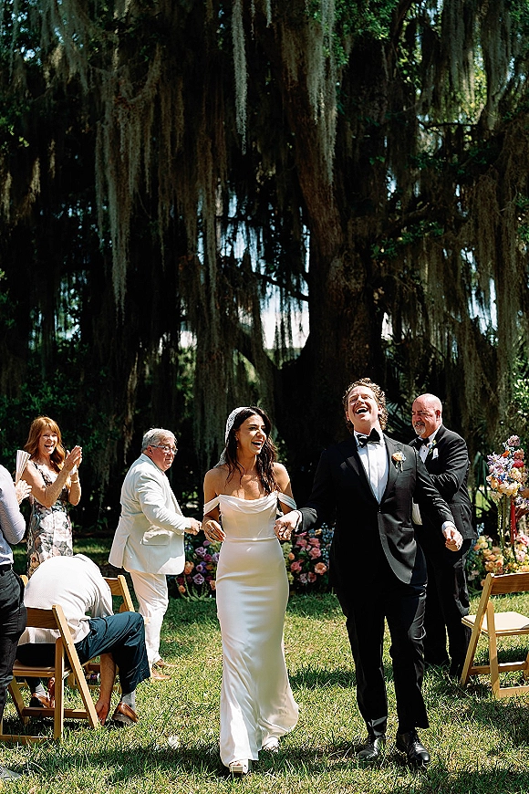 Wedding recessional as bride and groom walk the aisle holding hands, laughing in veil and tuxedo beneath an oak tree with hanging moss