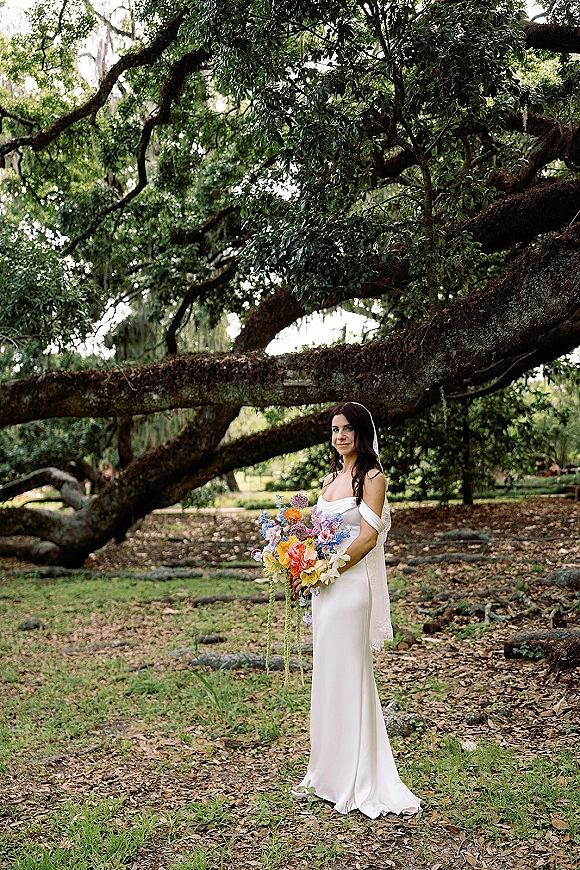 Bridal portrait of a bride holding bouquet in an off shoulder wedding dress under oak tree branches in a leafy park, veil trailing behind