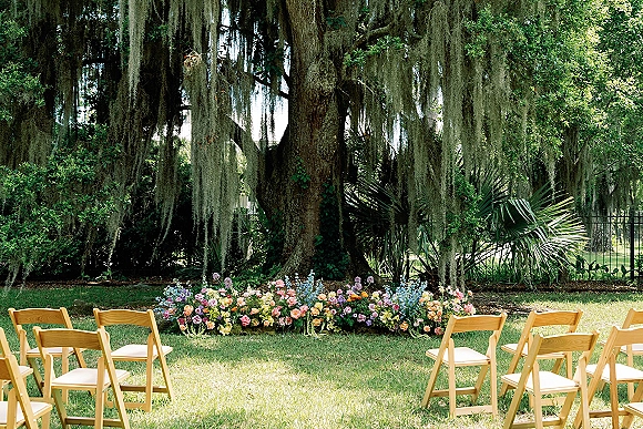 Ceremony setup for an outdoor wedding ceremony with wood folding chairs in a semicircle and a rose ground arrangement under an oak tree with Spanish moss