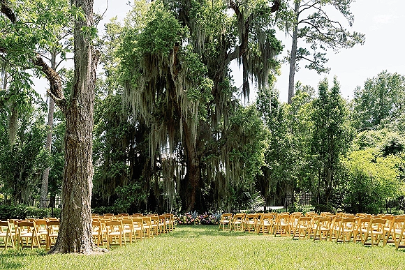 Outdoor ceremony setup with wood folding chairs and floral arrangements on a grass lawn beneath oak trees draped in moss by a black fence