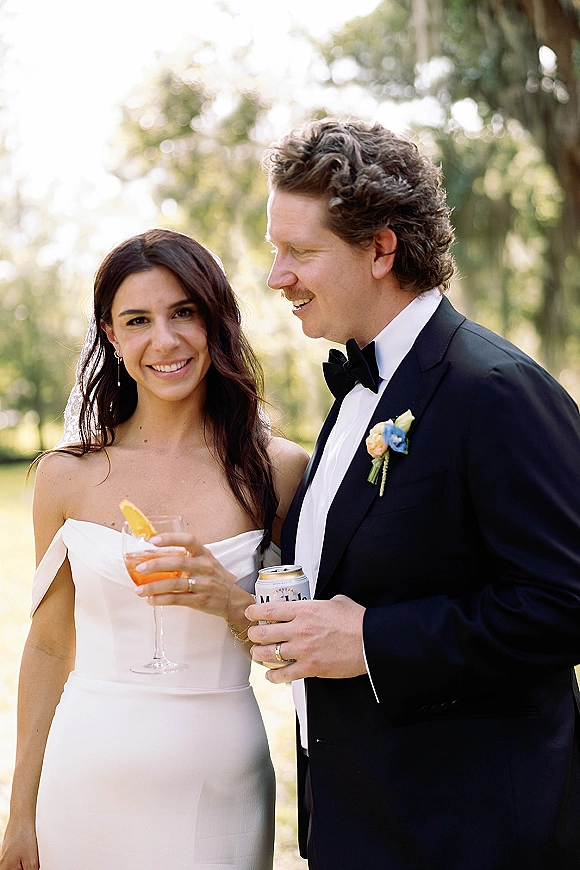 Couple portrait of bride and groom portrait smiling as she holds a cocktail with citrus garnish, in sunlit greenery with bokeh