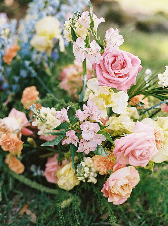 Wedding floral arrangement of pink roses and pastel blooms with greenery, styled on grass in an outdoor garden with soft bokeh backdrop