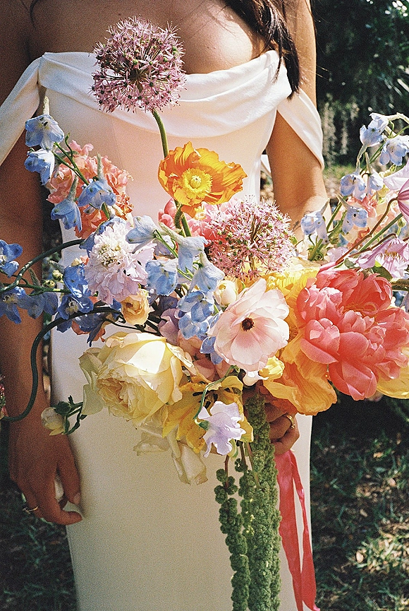 Bridal bouquet with colorful wedding bouquet blooms, trailing greenery and ribbon held against an off-the-shoulder dress in sunlit garden greenery