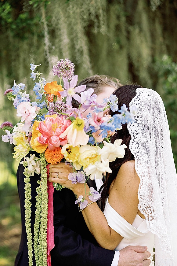 Wedding kiss portrait with a colorful wildflower bouquet covering the couple’s faces, lace veil and suit in a garden with mossy trees