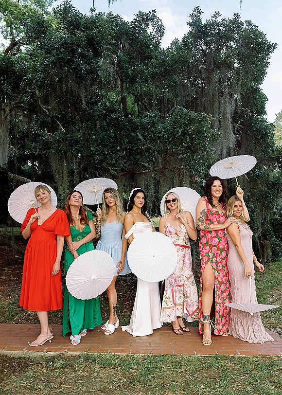 Bridesmaids group photo with white paper parasols, bride beside them in mismatched dresses under oak trees draped in moss on lawn