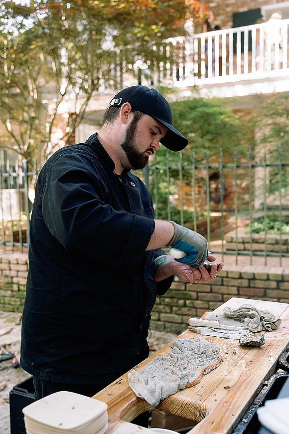 Oyster shucking at a wedding oyster bar as a chef in gloves uses an oyster knife at a wooden table on an outdoor patio