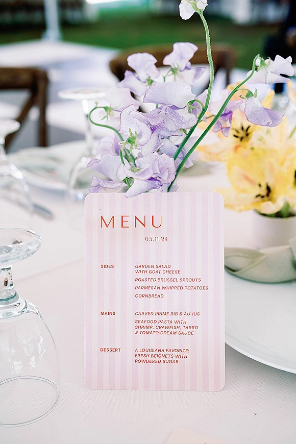Wedding menu card with pink stripes standing on a white tablecloth beside lilac and yellow flowers at an outdoor reception table setting