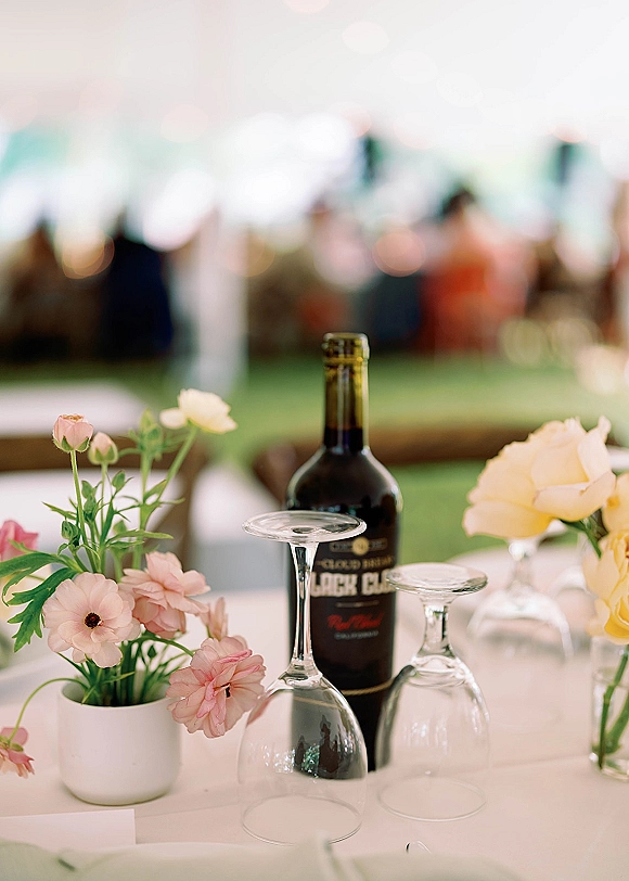 Reception tablescape with wine bottle table decor, upside down wine glasses, and pink flowers with yellow roses on a white tablecloth under a tent