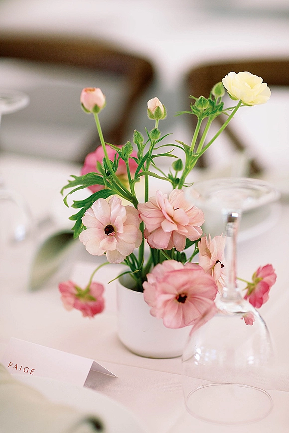 Wedding table centerpiece in a white bud vase with pink flowers beside a place card and wine glass on crisp linen, banquet chairs behind