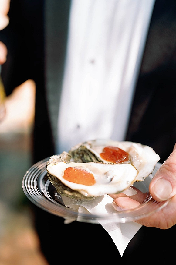 Oyster appetizer on a clear plate with cocktail sauce, held by a guest in a tuxedo jacket during an outdoor wedding cocktail hour with blurred guests