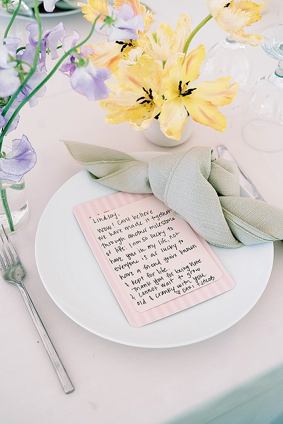 Wedding place setting with a white plate and sage green knotted napkin, handwritten note card, wine glass, and pastel bud vase flowers on a light tablecloth
