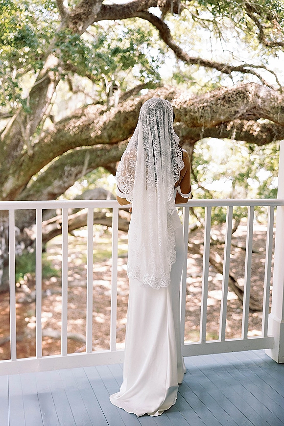 Bridal portrait of a back view bride in a white off-the-shoulder dress with a long lace veil, standing on a porch under tree branches