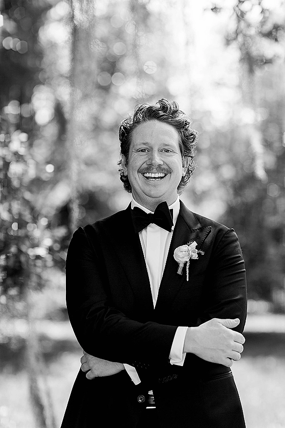 Groom portrait in a black tuxedo with bow tie and boutonniere, smiling outdoors against soft bokeh trees and greenery