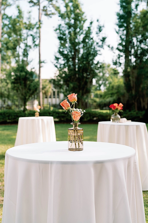Cocktail table decor with white linens and a bud vase of peach roses and baby's breath, set on a garden lawn with trees behind