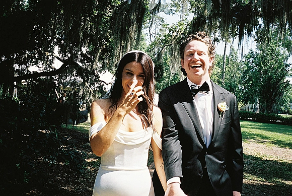 Couple portrait of bride and groom laughing hand in hand, bride in veil and satin dress, groom in tux, on a sunlit garden path with mossy trees
