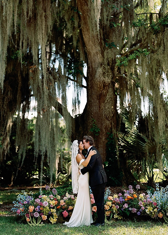 Wedding kiss portrait of bride and groom kissing beneath a moss-draped tree, veil and tux crisp in dappled garden sunlight
