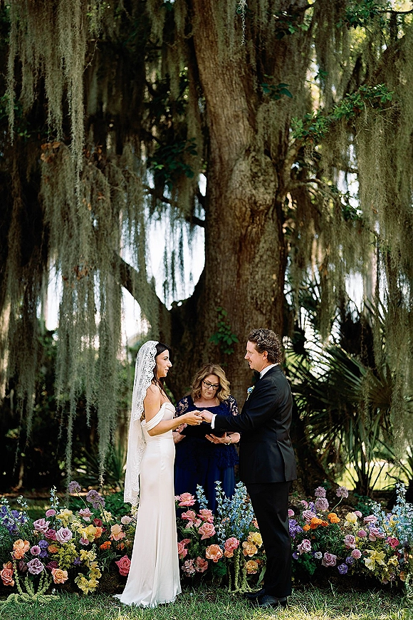 Wedding vows as bride and groom exchange rings during an outdoor wedding ceremony under an oak tree with spanish moss and floral ground arrangements