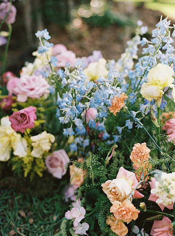 Wedding florals with pastel wedding flowers—roses, blue delphinium, carnations, and greenery arranged low against soft garden bokeh light