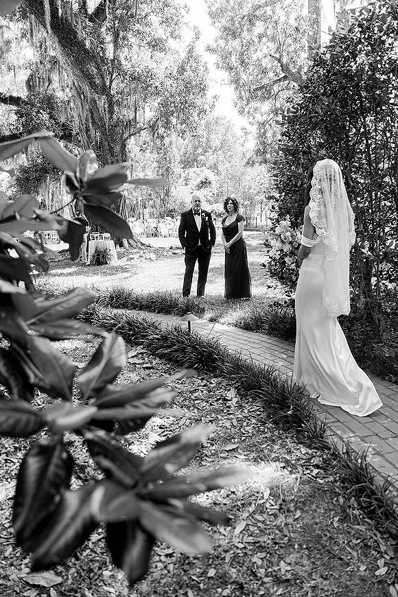 First look moment as bride approaches groom on a brick garden pathway, holding bouquet with lace veil, tuxedo and boutonniere under mossy trees