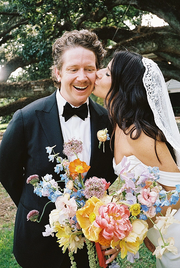 Wedding kiss portrait of bride kissing groom on the cheek, lace veil and colorful wildflower bouquet against a garden lawn and trees