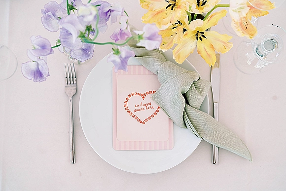 Wedding place setting with a sage green napkin, menu card and pastel flowers, including a yellow lily and sweet peas on light linen