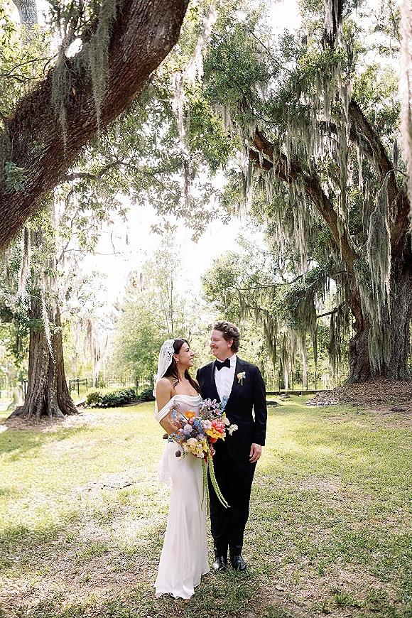 Couple portrait of bride and groom smiling, she holds a colorful bouquet with veil, under sunlit oak trees with hanging moss