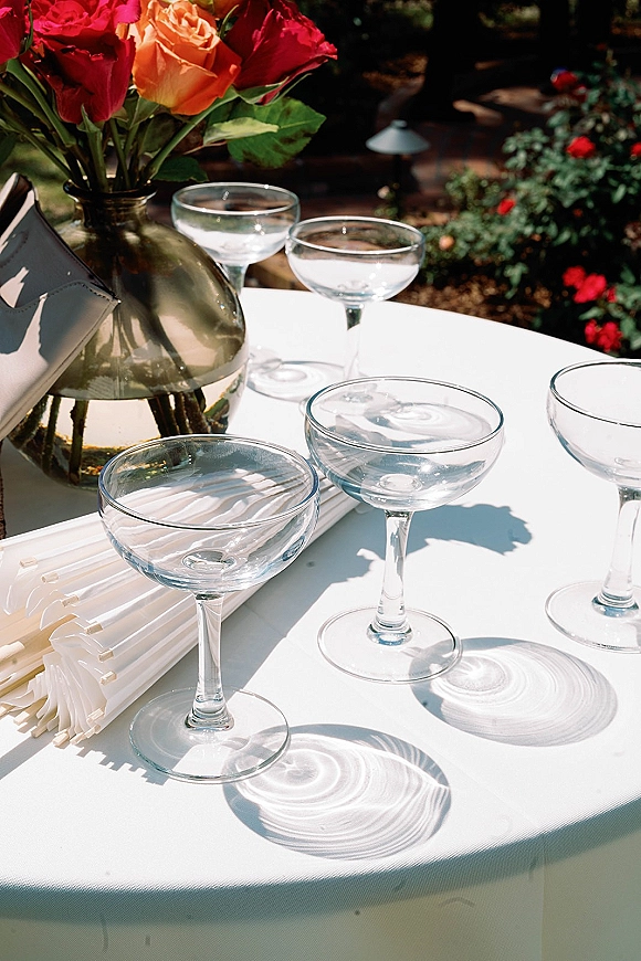 Reception tablescape with coupe glassware wedding place settings, rose centerpiece in glass vase on white tablecloth amid garden greenery outdoors